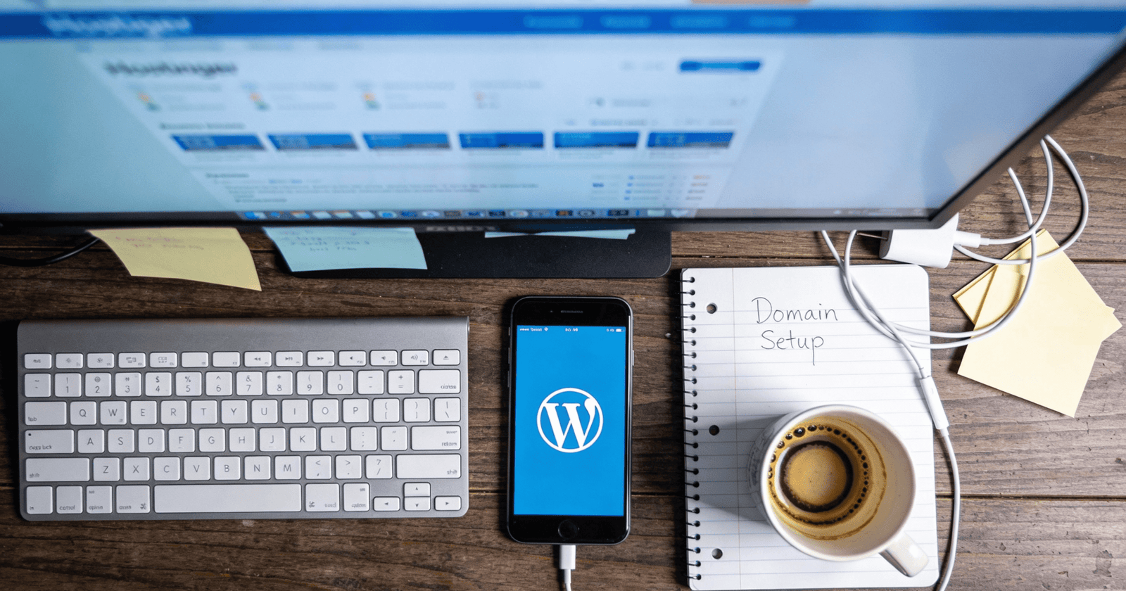 Overhead view of real blogger workspace with desktop monitor showing Hostinger dashboard, WordPress setup notes, smartphone, coffee mug, and planning materials in natural cool lighting